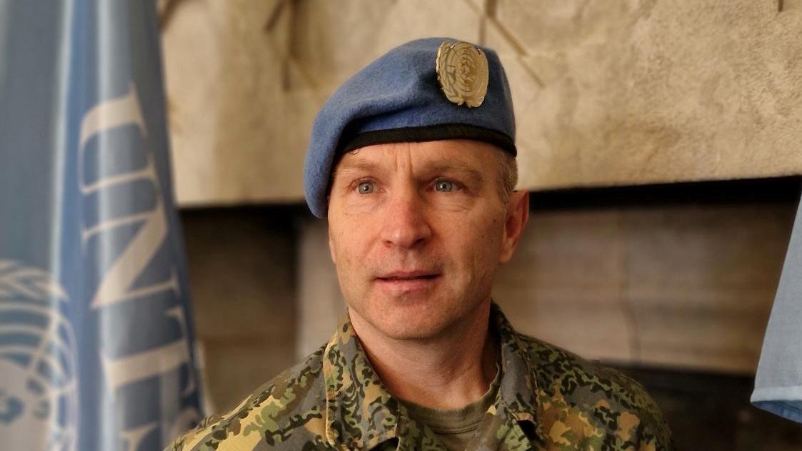 A man in military uniform and a blue beret poses for a photo in front of a UN flag 